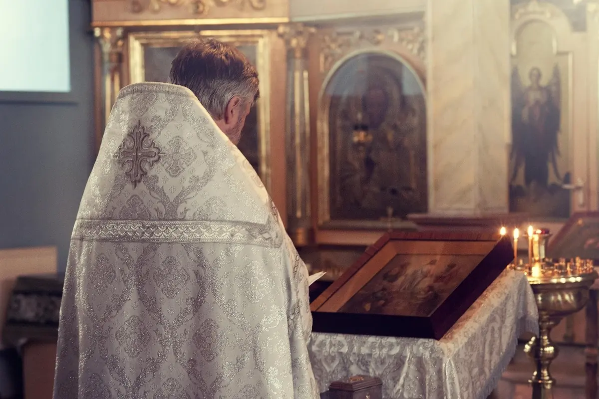 Священник у світлих ризах стоїть перед аналоєм з іконою, поруч горять свічки